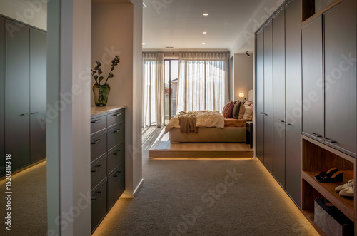 Photos A contemporary walk-in wardrobe leading to a stylish bedroom suite, featuring built-in cabinetry, LED strip lighting, and plush carpet flooring, with natural light filtering through sheer curtains