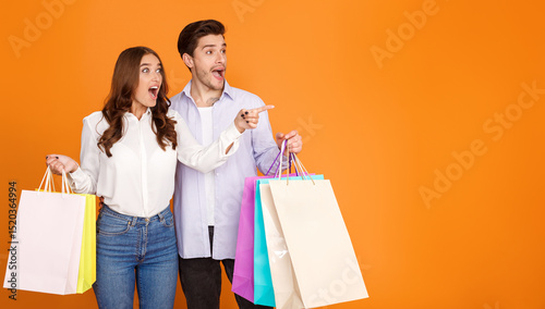 Wow sale. Shocked couple with shopping bags pointing fingers at free space, orange studio background photo