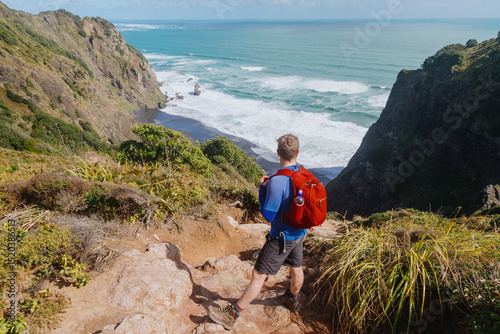 Hiker stands on a cliff overlooking a black sand beach and the Tasman Sea in New Zealand, enjoying the stunning coastal view. Mercer Bay, Karekare, Auckland, New Zealand