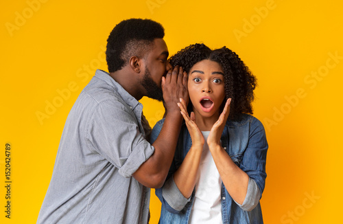 Hot Gossip. Black Guy Sharing Secret With His Shocked Girlfriend. Surprised Woman Opens Mouth In Disbelief Over Yellow Studio Background photo