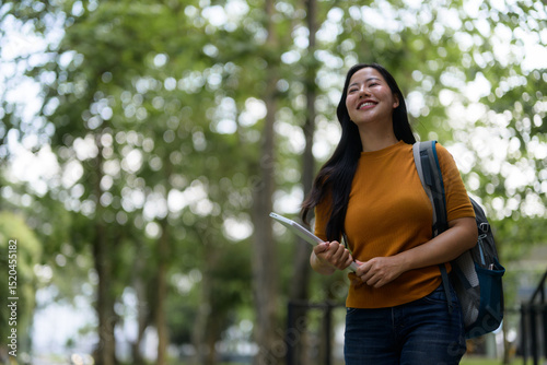 Happy asian student walking in university park holding tablet and backpack, enjoying campus life and looking up