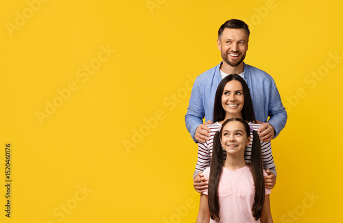 Happy Young Family, Mother Father And Daughter Embracing While Standing In Line, Cheerful Caucasian Parents And Teen Female Child Bonding Together, Posing On Yellow Studio Background, Copy Space