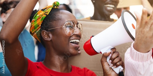 Smiling woman with megaphone, wearing glasses and colorful headscarf, leads a protest. Energetic woman, African descent, rallying with megaphone, inspiring others. African American woman protest.