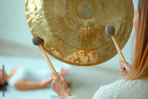 Gong bath therapy. Sound Healing Therapy. Session. A therapist strikes a golden gong during a sound healing session. A woman lies on a bed receiving the relaxing therapy. wellness, spa, meditation 