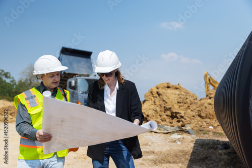 Two construction professionals examine site blueprints at an active urban construction area, with a dump truck, excavator, drainage pipes, and a tall building visible under a bright sky. photo