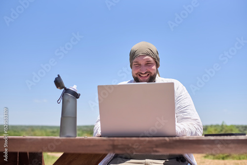 Young Hispanic man smiling while working on his laptop at an outdoor table on a sunny day. Concept: digital nomad working remotely while traveling enjoying a lifestyle of freedom.