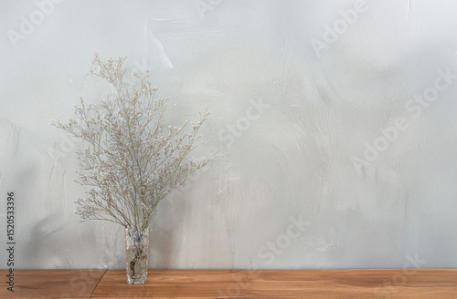 A minimalist arrangement of dried flowers in a clear vase on a wooden table, against a light gray wall.