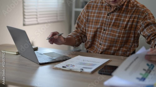 A man is sitting at a desk with a laptop and a stack of papers. He is using a pen to write on the papers