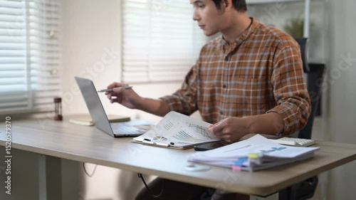 A man is sitting at a desk with a laptop and a stack of papers. He is looking at the papers and seems to be focused on the content. Concept of concentration and productivity