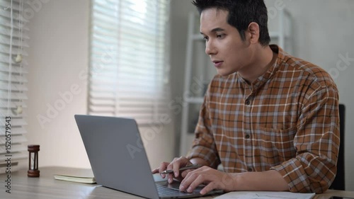 A man is sitting at a desk with a laptop open in front of him. He is typing on the keyboard and holding a pen in his hand