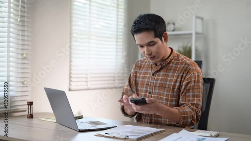 A man is sitting at a desk with a laptop and a stack of papers. He is typing on the laptop and he is focused on his work. The room has a modern and professional feel, with a chair