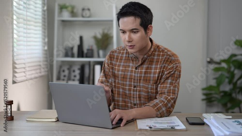A man is sitting at a desk with a laptop and a cell phone. He is typing on the laptop and looking at the cell phone. The scene suggests that he is working or studying