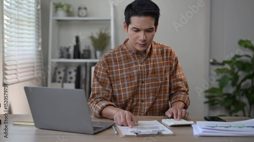 A man is sitting at a desk with a laptop and a keyboard. He is looking at a piece of paper and pointing at it. The scene suggests that he is working on a project or task that requires him to focus