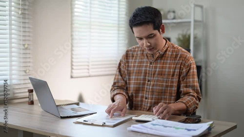 A man is sitting at a desk with a laptop and a clipboard. He is looking at the clipboard and pointing at something. The room has a window and a clock on the wall