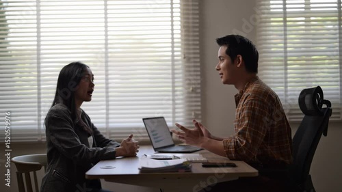 Two people are sitting at a table, one of them is talking to the other. The man is wearing a plaid shirt and the woman is wearing a gray jacket. They are both looking at a laptop
