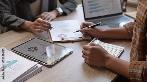 Two people are working on a project, one of them is writing on a tablet. The tablet is on a table with a calculator and a clipboard. The people are focused on their work