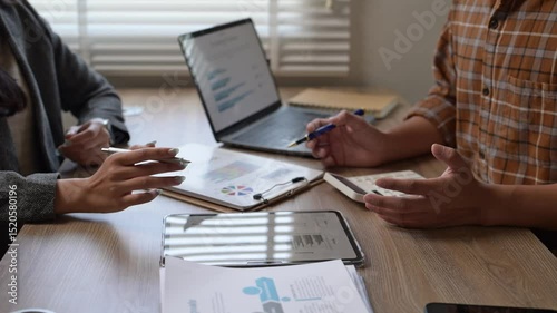 Two people are sitting at a table with laptops and papers. They are discussing something important. Scene is serious and focused
