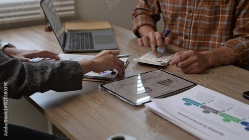 Two people are sitting at a table with a laptop, a calculator, and a notebook. They are discussing something important, possibly related to business or finance. The atmosphere seems serious