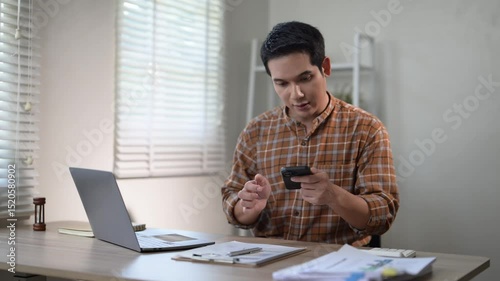 A man is sitting at a desk with a laptop and a clipboard. He is writing on the clipboard with a pen