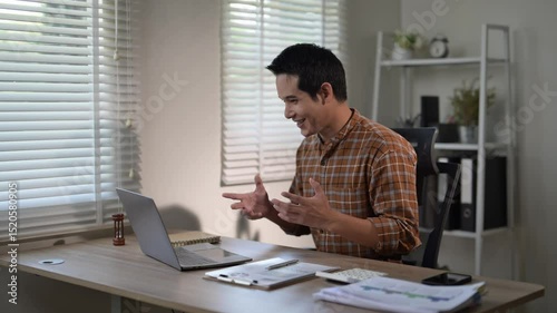 A man is sitting at a desk with a laptop and a stack of papers. He is wearing a plaid shirt and he is focused on his work