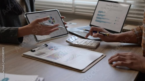 A group of people are working on a project, with one person pointing at a calculator and another person pointing at a tablet. The atmosphere seems to be focused and collaborative