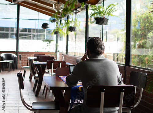 Photograph of an adult man talking on the phone in a cafe in front of a laptop at a wooden table with a textile tablecloth, with other tables covered in fabric, seen from behind. photo