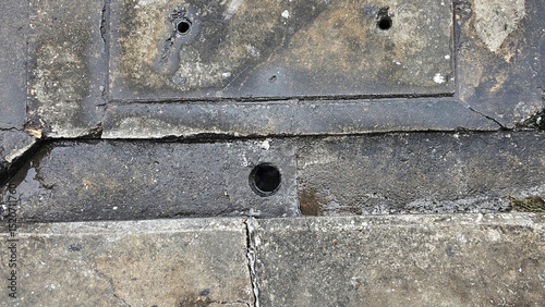 A close-up view of a weathered manhole cover on a gritty urban pavement, Weathered concrete sidewalk with a square manhole cover and visible stains, alongside a drainage gutter.	