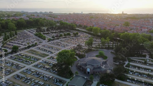 Aerial view of a large, neatly arranged cemetery with winding paths and gravestones, surrounded by trees and adjacent to a residential area under a clear blue sky.