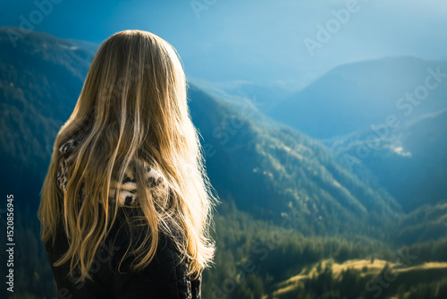 Young girl enjoying beauty of nature looking at mountains. Woman with long blonde hair standing on mountain top, admiring scenic valley and sunlit hills