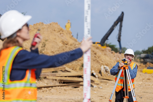 A construction surveyor uses a total station while another holds a leveling rod at a worksite filled with dirt and machinery, capturing accurate land measurements for site planning and layout. photo
