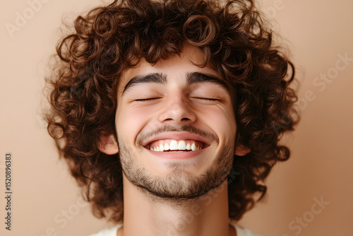 Portrait of a cheerful young man with curly hair laughing with closed eyes isolated over beige background photo