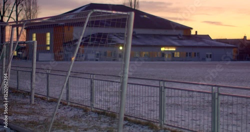 A snowcovered soccer field with a goalpost and a building in the sunset background, creating serenity