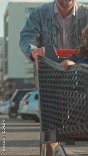 Vertical orientation. Excited boy riding inside shopping cart pushed by smiling father in parking lot, both enjoying playful moment. Child laughing during fun family grocery trip outdoors.