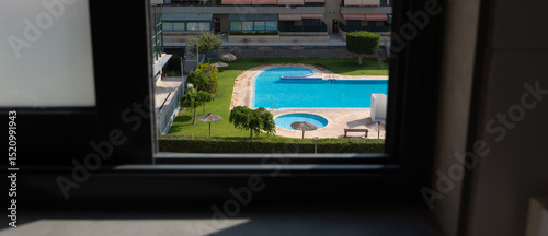 Framed view through a window shows a sparkling turquoise pool in a modern courtyard. Lush green lawn and clear blue water radiate summer vibes. Selective focus used.