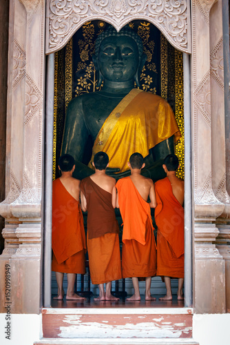 young buddhist novice monks, seen from behind, looking at big buddha statue in golden sash in Wat Chedi Luang temple, Chiang Mai, Thailand.