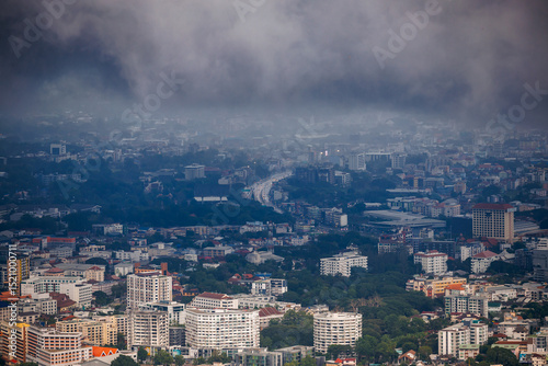 aerial view of chiang mai cityscape under a cloudy, rainy season sky, with urban buildings, a winding road, and green trees partially obscured by mist.
