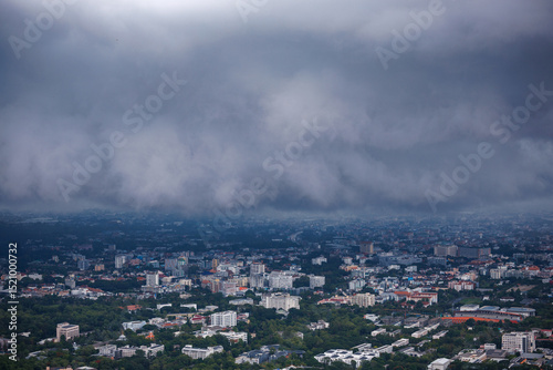 aerial view of chiang mai cityscape under a cloudy, rainy season sky, with urban buildings, a winding road, and green trees partially obscured by mist.
