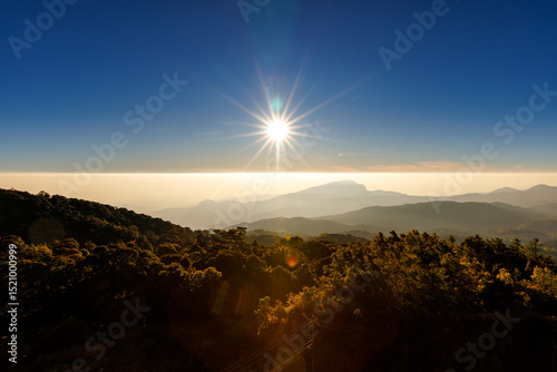  stunning wide shot captures a bright sun starburst over a hazy mountain range at sunrise or sunset, with dark forest treetops in the foreground and a clear blue sky above.