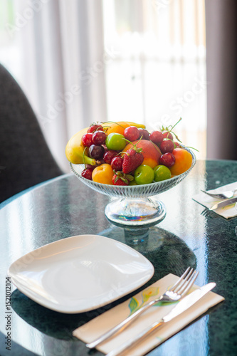 Softly lit secene of colorful fresh fruit bowl on marble tabletop with plates and chairs in the background in front of a window