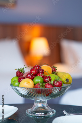 Colorful fresh fruit bowl on hotel room counter with armchair, bed and lampshade showing in the background
