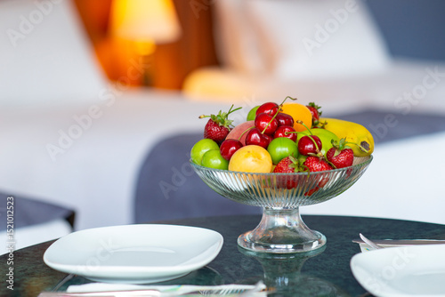 Colorful fresh fruit bowl on hotel room counter with armchair, bed and lampshade showing in the background