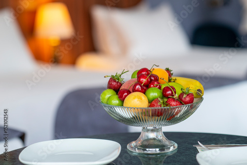 Colorful fresh fruit bowl on hotel room counter with armchair, bed and lampshade showing in the background