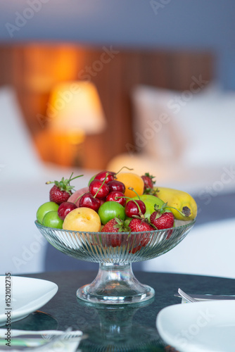 Colorful fresh fruit bowl on hotel room counter with armchair, bed and lampshade showing in the background