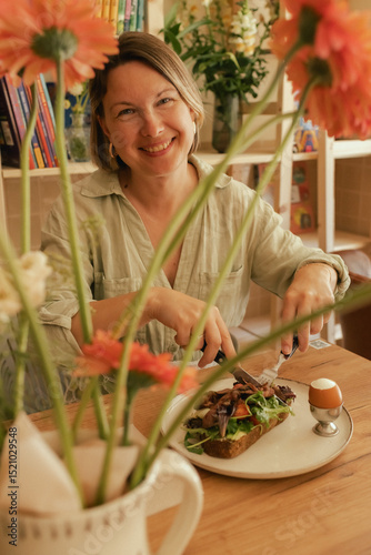 Crop middle-aged woman wearing stylish outfit eating delicious breakfast including egg and bacon with freshly greens served on wooden table in sunny outdoor cafe. Part of a series