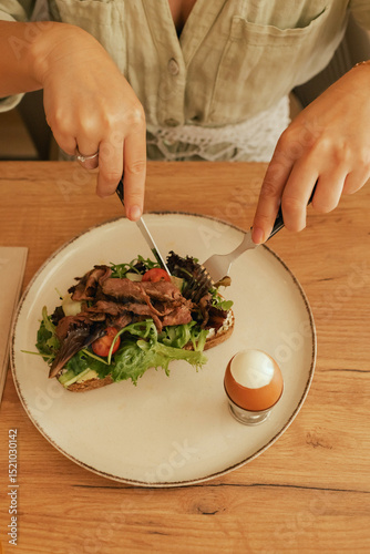 Close-up of female hands with cutlery having breakfast with egg and bacon with freshly greens served on wooden table in sunny outdoor cafe. Part of a series