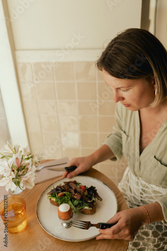 Crop middle-aged woman wearing stylish outfit eating delicious breakfast including egg and bacon with freshly greens served on wooden table in sunny outdoor cafe. Part of a series