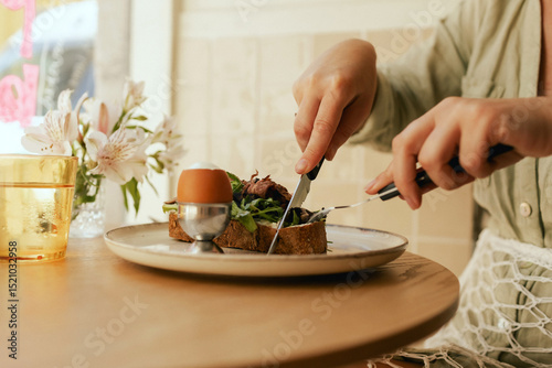 Close-up of female hands with cutlery having breakfast with egg and bacon with freshly greens served on wooden table in sunny outdoor cafe. Part of a series