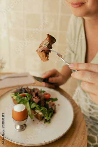 Close-up of female hands with cutlery having breakfast with egg and bacon with freshly greens served on wooden table in sunny outdoor cafe. Part of a series