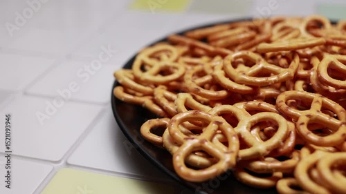 Camera Moves From Left To Right, Revealing A Black Plate Piled High With Golden Brown Salty Pretzel Snacks. Appetizing Food Close Up.