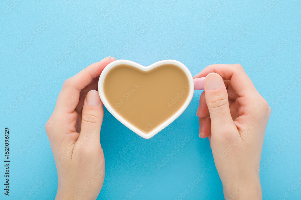 Obraz premium Young adult woman hands holding white heart shape cup of fresh coffee with milk on light blue table background. Pastel color. Closeup. Point of view shot. Top down view.